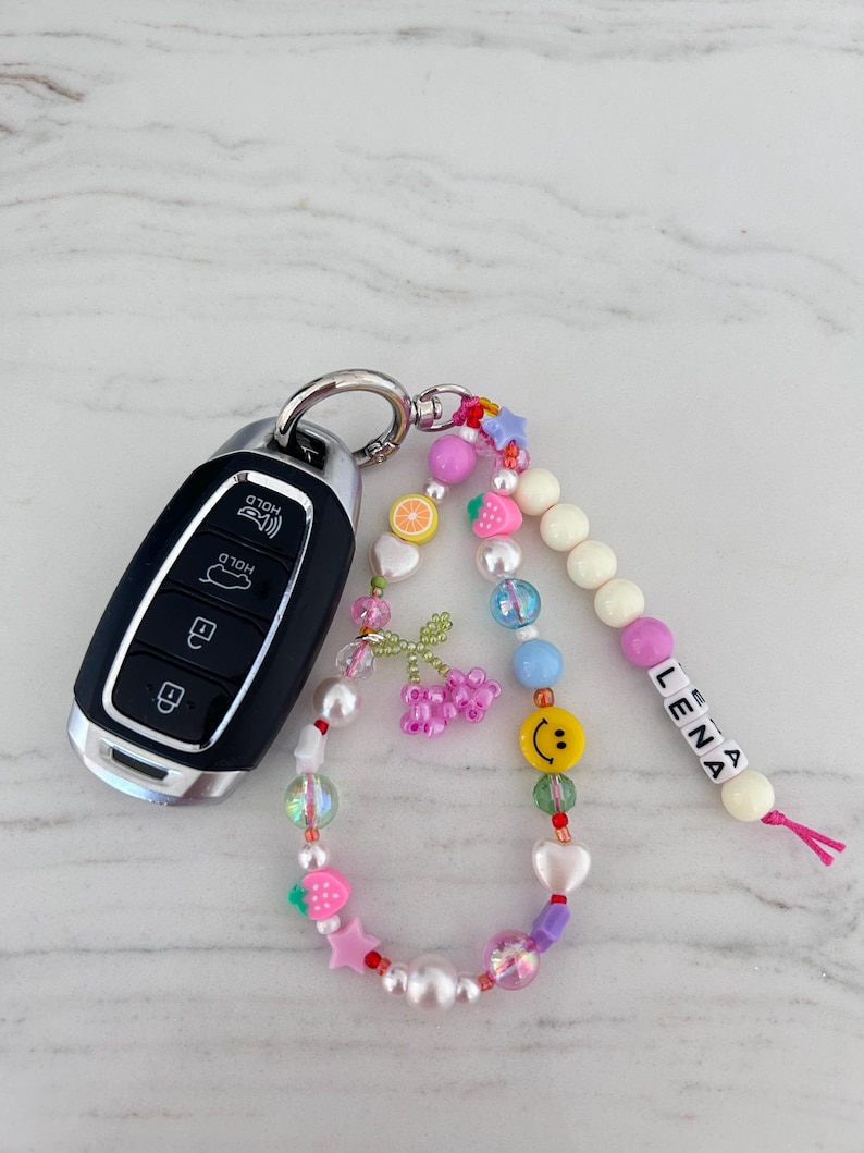 May include: A colorful beaded keychain with a smiley face charm, fruit charms, and a name tag that reads "LENA". The keychain is attached to a car key.