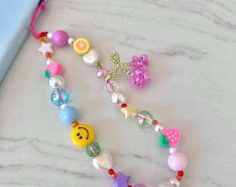 May include: A colorful phone charm with a variety of beads, including white pearls, a smiley face, a star, a strawberry, a lemon, and a cluster of pink beads. The charm is attached to a light blue phone case.