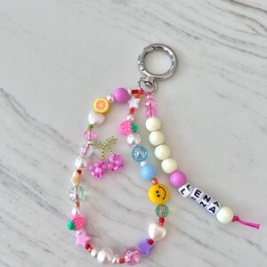 May include: A colorful beaded keychain with a silver key ring. The beads are pink, white, green, blue, yellow, and red. The keychain has a charm with a smiley face, a charm with a strawberry, and a charm with a cherry. The keychain also has a charm with the name "ELENA" spelled out in white letters.