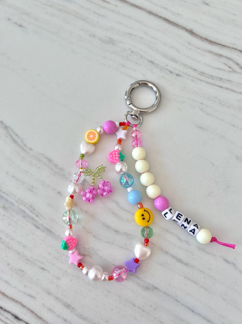 May include: A colorful beaded keychain with a silver key ring. The beads are pink, white, green, blue, yellow, and red. The keychain has a charm with a smiley face, a charm with a strawberry, and a charm with a cherry. The keychain also has a charm with the name "ELENA" spelled out in white letters.