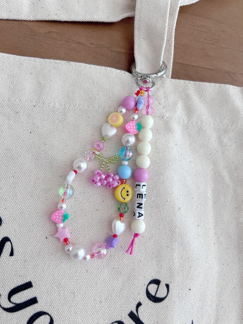May include: A colorful beaded keychain with a white background. The keychain features a variety of charms, including strawberries, stars, hearts, and a smiley face. The keychain also includes the name "Lena" spelled out in white letters.