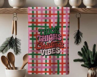 Puede incluir: Un paño de cocina a cuadros rosa, rojo y verde con el texto "THICK THIGHS & CHRISTMAS VIBES" en una fuente retro. El paño está colgado en un estante de madera con otros artículos de cocina y decoración navideña.