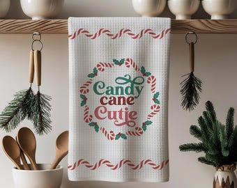 Puede incluir: Toalla de cocina blanca con tejido de gofre y diseño de bastones de caramelo rojos y verdes. La toalla presenta el texto "Candy cane Cutie" en verde y rojo, rodeado por una corona de bastones de caramelo. Accesorio de cocina festivo.