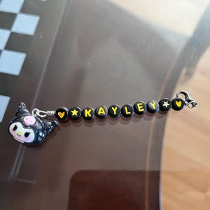 May include: Black beaded bracelet with a charm of a black and white cartoon character with pink accents. The beads are black with yellow letters spelling out the name "KAYLEY" and yellow heart and star charms.