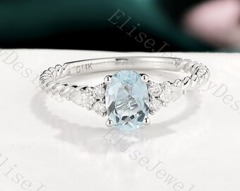 May include: A delicate silver ring with a twisted band and a light blue oval gemstone surrounded by smaller diamonds. The ring is set on a white background.