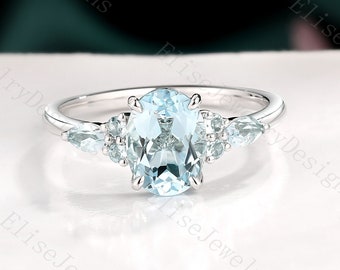 May include: A delicate oval aquamarine engagement ring set in white gold with smaller accent stones. The ring features a light blue aquamarine center stone and smaller clear stones on either side.