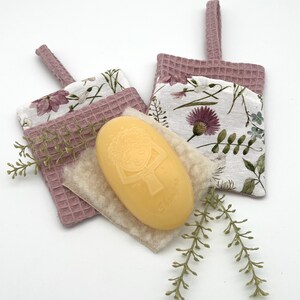 Könnte beinhalten: Zwei mauvefarbene Seifenhalter aus Waffelstoff mit floralem Innenfutter und Aufhängeschlaufen. Eine hellgelbe ovale Seife mit einem Kindergesicht-Design liegt auf einem beigen Waschlappen. Grüne Zweige sorgen für eine dekorative Note.