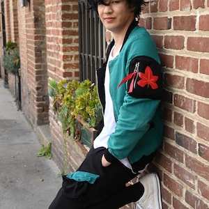 May include: A young person wearing a teal green bomber jacket with a black patch featuring a red maple leaf design. The jacket has a white zipper and a white t-shirt underneath. The person is wearing black pants with a teal green pocket and white sneakers.