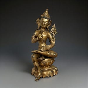 May include: A golden statue of a seated figure in a meditative pose. The figure wears a crown and ornate jewelry, with one hand raised in a gesture of blessing. The statue is seated on a lotus flower base, with intricate details throughout.
