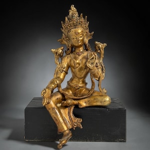 May include: A golden statue of a seated deity with ornate headdress and jewelry. The figure is posed with one leg crossed and the other extended. The statue is mounted on a black rectangular base. The overall style suggests a religious or spiritual theme.