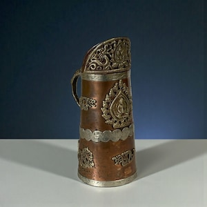 Puede incluir: Jarra decorativa de cobre y plata con detalles intrincados. La jarra presenta un asa, un borde decorativo y adornos en relieve plateados y dorados, incluida una figura central de Buda.