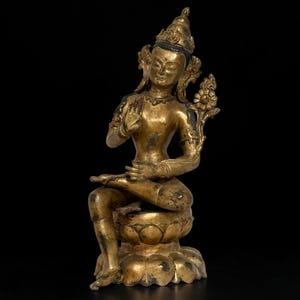 May include: A gold-toned metal statue of a seated figure in a meditative pose, resting on a lotus-shaped base. The figure features an ornate crown, jewelry, and a floral element extending from the left shoulder, with one hand raised in a gesture.