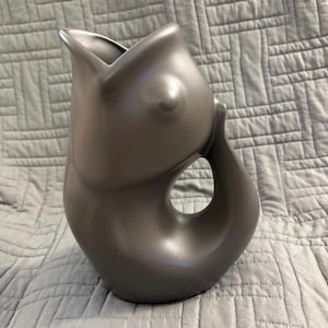 May include: A matte dark gray ceramic pitcher shaped like a fish with an open mouth at the top, a protruding eye, and a curved tail section that forms a handle. The pitcher has a smooth, solid-colored finish and a distinct aquatic silhouette.