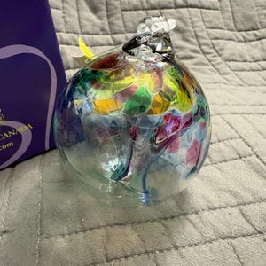 May include: A spherical glass ornament featuring a clear base with internal swirls of multicoloured glass in shades of blue, green, yellow, and pink. The top of the ornament has a clear glass loop for hanging, and the surface is smooth and reflective.