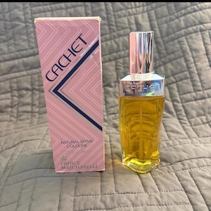 Könnte beinhalten: Eine rechteckige rosa Schachtel mit weißen geometrischen Linien und dem Text CACHET sowie PRINCE MATCHABELLI, neben einem durchsichtigen Kölnischwasser-Flakon aus Glas mit silbernem Verschluss und goldener Flüssigkeit, mit Prägung CACHET am Kragen.