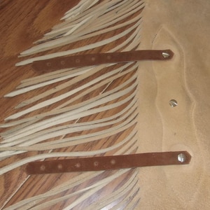 May include: Close-up of a tan leather item with long, cream-colored fringe. Two brown leather straps with rivets are visible. The item appears to be a piece of clothing or accessory, possibly a vest or chaps.