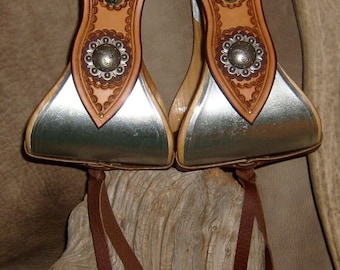 May include: A pair of brown leather stirrup leathers with silver conchos and metal stirrups. The leathers are attached to a brown leather strap.