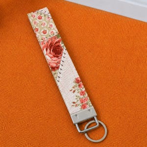 May include: A floral fabric wristlet key fob with a metal key ring. The key fob features a patchwork design with a large red rose and smaller rose accents on a cream background. The key fob is approximately 6 inches long.