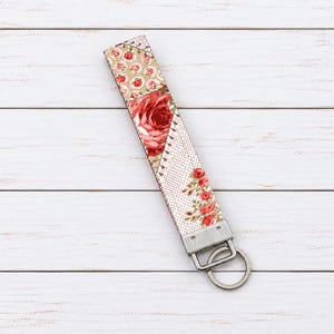 May include: A fabric wristlet keychain featuring a patchwork design with red roses on a beige background, a large central red rose, and a section with small red floral patterns on a white dotted background, finished with a silver metal clasp.
