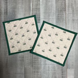 May include: Two square quilted pot holders featuring a cream-colored fabric with a repeating pattern of small green rabbits and tiny dots. Each pot holder is finished with a solid dark green fabric border stitched along all four edges.