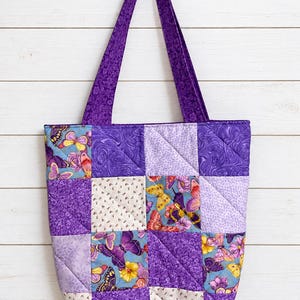 May include: A quilted tote bag featuring a patchwork design of purple, lavender, and white fabric squares. The pattern includes various floral and butterfly prints in shades of yellow, pink, and blue. It has two long purple fabric shoulder straps.