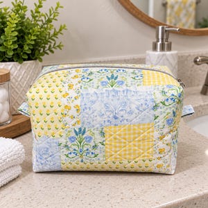 May include: A quilted fabric pouch featuring a patchwork design of yellow and blue floral patterns, small yellow flower prints, and yellow gingham checks. The rectangular bag has a silver-toned metal zipper closure along the top edge.