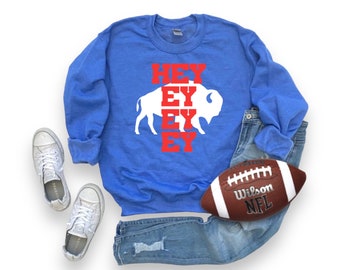 Puede incluir: Sudadera azul con las palabras "HEY EY EY EY" en rojo y un gráfico de búfalo blanco. La imagen incluye vaqueros azules, zapatillas blancas y un balón de fútbol americano marrón y blanco.