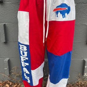 May include: Red, white, and blue sweatpants with the word "BUFFALO" in white block letters down the left leg. The right leg features a blue buffalo logo. The pants have a white drawstring and waistband.