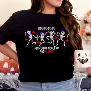 May include: Black t-shirt featuring a graphic of dancing skeletons with red, white, and blue accents. The text on the shirt reads "HEY-EY-EY-EY" and "KICK YOUR HEELS UP AND SHOUT!" A Halloween-themed design.