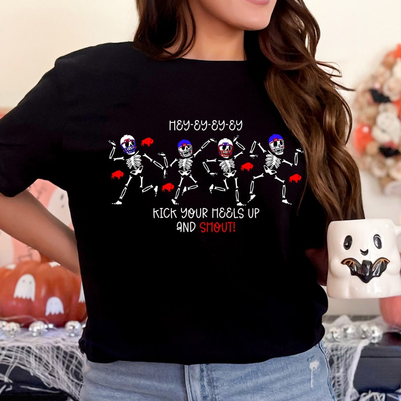 May include: Black t-shirt featuring a graphic of dancing skeletons with red, white, and blue accents. The text on the shirt reads "HEY-EY-EY-EY" and "KICK YOUR HEELS UP AND SHOUT!" A Halloween-themed design.
