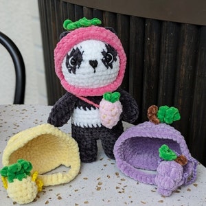 May include: A black and white crocheted panda bear wearing a pink crocheted strawberry hat. The panda is holding a crocheted strawberry. There are two crocheted fruit hats, one yellow pineapple and one purple grape, on the table.