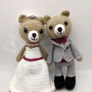 May include: A crocheted wedding couple bear set. The bride bear is wearing a white dress with a burgundy sash and a veil. The groom bear is wearing a gray suit with a burgundy bow tie.