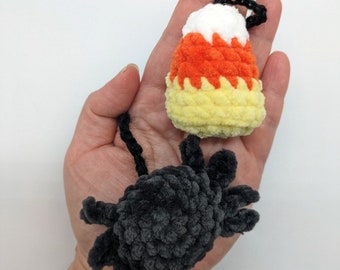 May include: A hand holding two crocheted Halloween decorations. One is a black spider and the other is a candy corn.