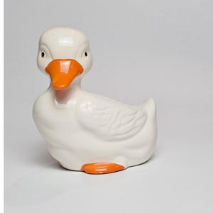 May include: A white ceramic duck ornament with an orange beak and feet. The duck is depicted in a sitting pose and has a glossy finish. This decorative item is likely for display.