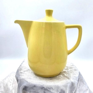 Puede incluir: Una tetera de cerámica amarilla vintage con tapa y asa. La tetera tiene un cuerpo redondeado, un pico y un asa. La tapa tiene un pequeño pomo en la parte superior. La tetera está sobre una superficie blanca.