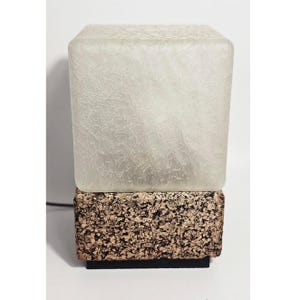 May include: A contemporary table lamp featuring a square, frosted glass shade atop a textured, cork-style base. The lamp has a black base and a power cord. The shade has a crackled effect, and the base is speckled brown and tan.