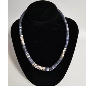 Op de afbeelding: Een ketting van cilindrische kralen, tentoongesteld op een zwarte mannequin. De kralen zijn donkerblauw en wit, gerangschikt in een herhalend patroon. Secties met witte kralen zijn verspreid. Dit is een sieraad.