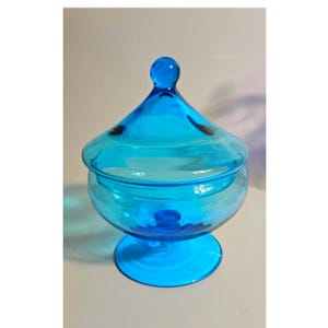 May include: A vibrant turquoise blue glass candy dish featuring a wide, rounded bowl, a pedestal base, and a matching removable lid with a spherical knob handle. The translucent glass construction allows light to pass through the entire vessel.