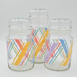 May include: Three clear glass jars with matching lids. Each jar is decorated with a colorful crisscross pattern of red, orange, yellow, green, blue, and purple lines. The jars are likely for kitchen storage.