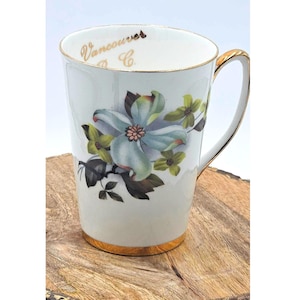 May include: White ceramic mug with a gold handle and rim, featuring a floral design in shades of blue, green, and brown. The mug has the text "Vancouver & Co." in gold lettering near the rim. The mug is on a wooden surface.