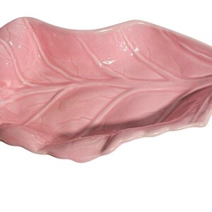 May include: A glossy, light pink ceramic dish shaped like a leaf. The dish features a smooth, curved surface with embossed vein details that mimic the natural structure of a leaf, including a pointed tip and scalloped, wavy edges throughout.