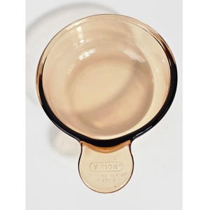 May include: A clear, amber-colored Vision Corning U.S.A. V-150-B saucepan with a black rim and handle. The handle has the Vision logo and product information. The saucepan is empty and appears to be made of glass.