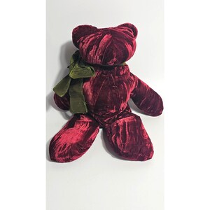 May include: A plush teddy bear crafted from deep burgundy velvet fabric. The bear features a rounded head, two small ears, and outstretched arms and legs. An olive green ribbon is tied in a bow around the neck of the stuffed animal.