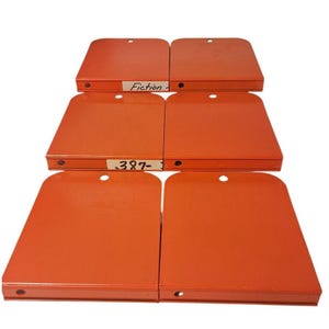 May include: Six orange metal library shelf dividers arranged in three rows of two. Each divider features a rounded top edge with a circular hole and a flat base with a small hole. Two dividers have white labels with the text Fiction and 387-.
