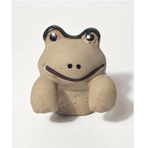 May include: A small, light beige ceramic frog figurine with a smooth, matte finish. The frog features a dark green painted top section, two black painted eyes, and a thin, dark horizontal line representing a mouth. The figure has two front limbs.