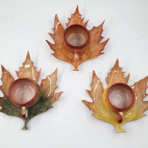 Puede incluir: Tres tazas de té de cerámica con platillos en forma de hoja. Las tazas son marrones y tienen un acabado brillante. Los platillos están decorados con un diseño de hojas en tonos marrones, amarillos y verdes.