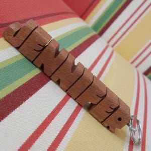 May include: A brown wooden keychain featuring the name JENNIFER carved in block letters. The letters are connected in a single horizontal piece, and a silver-colored metal split ring is attached to the end of the final letter R.