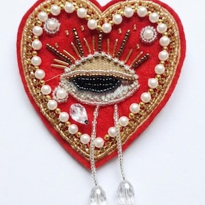 May include: A heart-shaped, red felt brooch with an embroidered eye design. The eye is detailed with gold, black, and silver thread. The brooch is embellished with pearls and gold beads, and has two dangling crystal pendants.