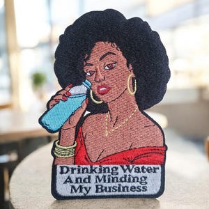 May include: Embroidered patch featuring a Black woman with large black hair, wearing gold hoop earrings and a red top, drinking from a blue water bottle. The patch includes the text "Drinking Water And Minding My Business."