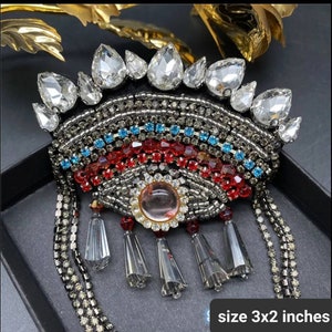 May include: A large, eye-shaped brooch with a beaded design. The brooch is made of black, red, blue, and silver beads. It has a large, clear crystal in the center and is surrounded by smaller crystals. The brooch is 3 inches by 2 inches.